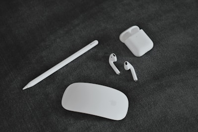 A collection of Apple gadgets, including a white stylus, a pair of wireless earbuds with their charging case, and a sleek, white computer mouse laid out on a dark, textured surface.