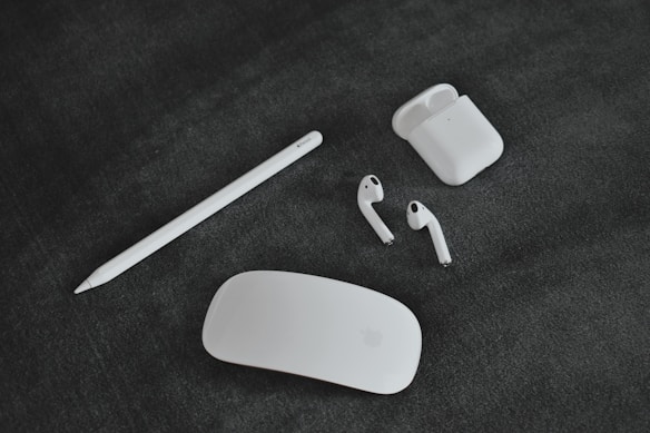 A collection of Apple gadgets, including a white stylus, a pair of wireless earbuds with their charging case, and a sleek, white computer mouse laid out on a dark, textured surface.