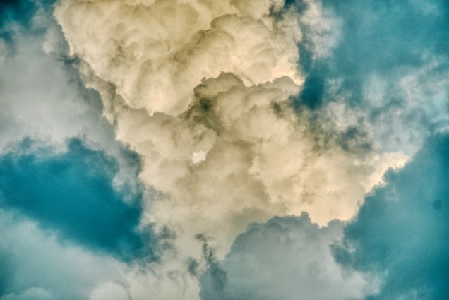 Billowing, fluffy clouds dominate the scene, with shades of cream and ivory contrasted against a background of blue sky. The texture of the clouds is soft and voluminous, creating a dynamic and natural pattern across the sky.