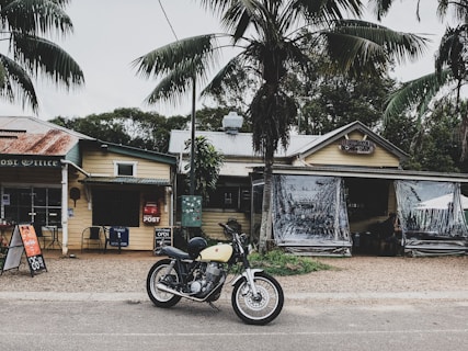 A vintage motorcycle is parked in front of an old rustic post office and cafe. The buildings have a weathered appearance with corrugated metal roofs and wooden structures. Palm trees surround the area, and there are signs for open hours and tourist information.