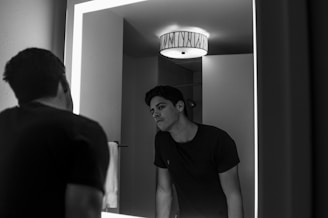 A person is standing in front of a mirror, leaning slightly forward, looking at their reflection. The scene is set in a bathroom with a circular ceiling light and a towel visible next to the mirror. The person is wearing a dark t-shirt.