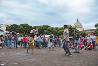 A lively street performance is taking place, where a young man is doing a handstand in the center of a crowd. The audience forms a circle around the performer, showing interest and engagement. In the background, there are green trees and a building with a dome, suggesting an outdoor public space.
