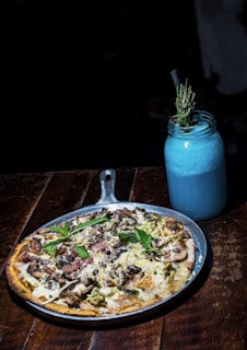 A delicious-looking pizza topped with various ingredients like cheese, mushrooms, and fresh basil leaves is served on a metal pan. Next to it is a blue drink in a mason jar with a straw, garnished with a sprig of rosemary. The setup is on a wooden table, creating a rustic and cozy atmosphere.