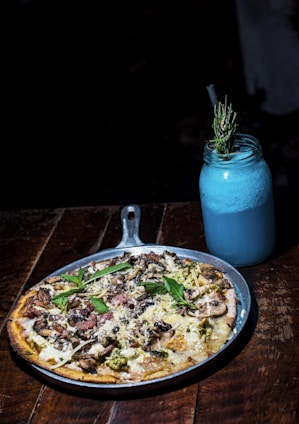 A delicious-looking pizza topped with various ingredients like cheese, mushrooms, and fresh basil leaves is served on a metal pan. Next to it is a blue drink in a mason jar with a straw, garnished with a sprig of rosemary. The setup is on a wooden table, creating a rustic and cozy atmosphere.