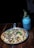 A delicious-looking pizza topped with various ingredients like cheese, mushrooms, and fresh basil leaves is served on a metal pan. Next to it is a blue drink in a mason jar with a straw, garnished with a sprig of rosemary. The setup is on a wooden table, creating a rustic and cozy atmosphere.