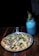 A delicious-looking pizza topped with various ingredients like cheese, mushrooms, and fresh basil leaves is served on a metal pan. Next to it is a blue drink in a mason jar with a straw, garnished with a sprig of rosemary. The setup is on a wooden table, creating a rustic and cozy atmosphere.