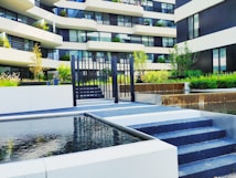 A modern residential building with multiple stories is visible, featuring a facade of white and dark panels. The foreground includes a neatly landscaped area with small trees and shrubs. A water feature with cascading water adds an elegant touch. Black metal railings and a set of steps lead to a gated area.