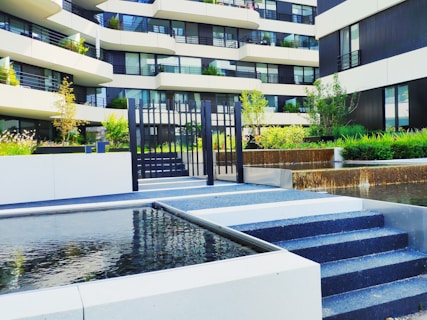 A modern residential building with multiple stories is visible, featuring a facade of white and dark panels. The foreground includes a neatly landscaped area with small trees and shrubs. A water feature with cascading water adds an elegant touch. Black metal railings and a set of steps lead to a gated area.