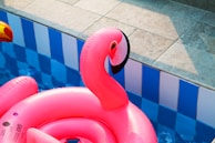 A large, bright pink inflatable flamingo float is positioned in a swimming pool, with the pool's blue water and patterned blue and white walls visible. The sun casts shadows across the poolside tiles, enhancing the vibrant colors of the scene.
