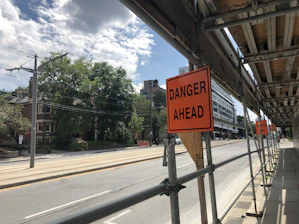 A city street is under construction, with scaffolding on the right side. There are multiple orange warning signs indicating 'DANGER AHEAD'. The road appears empty, with trees and residential buildings on the left side. Overhead, the sky is partly cloudy.