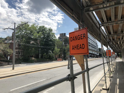 A city street is under construction, with scaffolding on the right side. There are multiple orange warning signs indicating 'DANGER AHEAD'. The road appears empty, with trees and residential buildings on the left side. Overhead, the sky is partly cloudy.