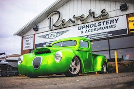 A custom bright green vintage truck is parked in front of a building with a sign advertising upholstery and accessories. The truck features a sleek, modified design with a prominent front grille and modern wheels. The background includes signage for window tint services and other automotive customizations.
