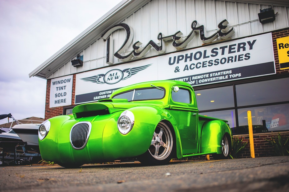 A custom bright green vintage truck is parked in front of a building with a sign advertising upholstery and accessories. The truck features a sleek, modified design with a prominent front grille and modern wheels. The background includes signage for window tint services and other automotive customizations.