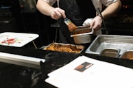 A person wearing a black apron is applying sauce with a brush onto cooked pieces of meat. The setting appears to be a kitchen, with various trays and a decorative plate featuring a floral design visible on the counter.