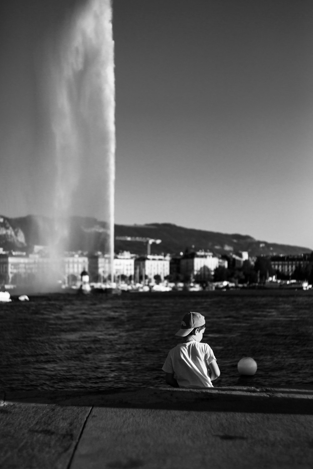 Photographie en niveaux de gris d’un garçon assis à côté d’un plan d’eau