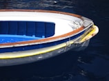 A small boat with a white and blue interior and brown trim floats on dark blue water. A coil of rope is seen at the front of the boat, indicating it may be used for docking. The hull features a yellow stripe along its edge, showing signs of wear.
