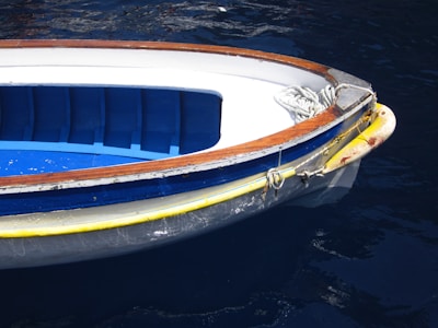 A small boat with a white and blue interior and brown trim floats on dark blue water. A coil of rope is seen at the front of the boat, indicating it may be used for docking. The hull features a yellow stripe along its edge, showing signs of wear.