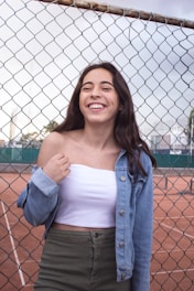 A young woman with long dark hair is smiling joyfully. She wears a white tube top, an open blue denim jacket, and green high-waisted trousers. She stands in front of a chain-link fence, with a clay tennis court visible behind her and a cloudy sky above.