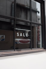 A window with black framing has the word 'SALE' written in bold, white capital letters on its glass. The background shows a blurred interior and exterior environment, possibly of a store or commercial building.