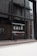 A window with black framing has the word 'SALE' written in bold, white capital letters on its glass. The background shows a blurred interior and exterior environment, possibly of a store or commercial building.