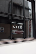 A window with black framing has the word 'SALE' written in bold, white capital letters on its glass. The background shows a blurred interior and exterior environment, possibly of a store or commercial building.