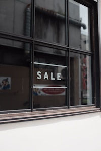 A window with black framing has the word 'SALE' written in bold, white capital letters on its glass. The background shows a blurred interior and exterior environment, possibly of a store or commercial building.