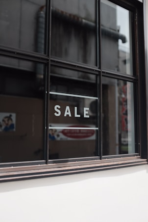 A window with black framing has the word 'SALE' written in bold, white capital letters on its glass. The background shows a blurred interior and exterior environment, possibly of a store or commercial building.