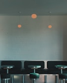 A minimalist interior features a row of black cushioned seating against a light brick wall. Three round pendant lights hang from the ceiling above small metal tables, creating a modern and cozy atmosphere.