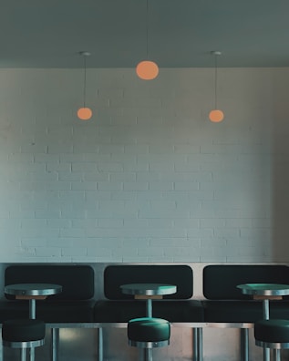 A minimalist interior features a row of black cushioned seating against a light brick wall. Three round pendant lights hang from the ceiling above small metal tables, creating a modern and cozy atmosphere.
