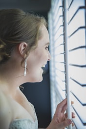 A graceful woman wearing soft champagne gold earrings, framed by sunlight filtering through sheer curtains.