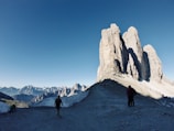 The image features a breathtaking mountain landscape with large, jagged peaks dominating the horizon under a clear blue sky. The foreground shows a rocky path where several hikers, equipped with trekking poles, are walking towards the towering rock formations. Snow patches are visible around the path, adding a stark contrast to the rugged terrain.