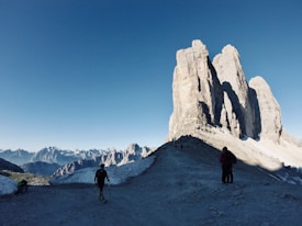 The image features a breathtaking mountain landscape with large, jagged peaks dominating the horizon under a clear blue sky. The foreground shows a rocky path where several hikers, equipped with trekking poles, are walking towards the towering rock formations. Snow patches are visible around the path, adding a stark contrast to the rugged terrain.