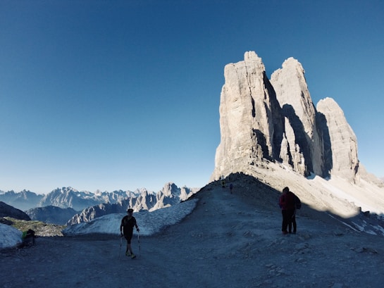 The image features a breathtaking mountain landscape with large, jagged peaks dominating the horizon under a clear blue sky. The foreground shows a rocky path where several hikers, equipped with trekking poles, are walking towards the towering rock formations. Snow patches are visible around the path, adding a stark contrast to the rugged terrain.