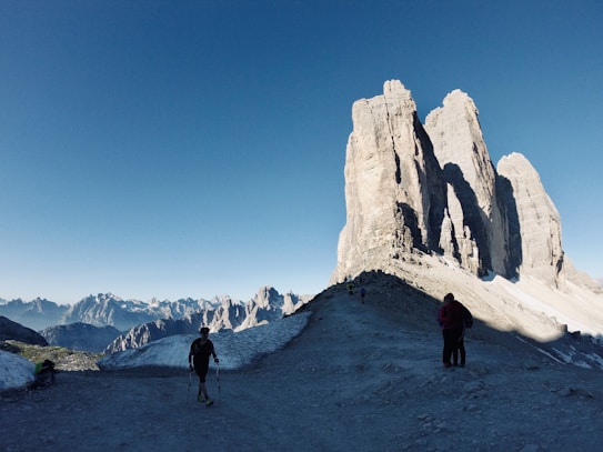 The image features a breathtaking mountain landscape with large, jagged peaks dominating the horizon under a clear blue sky. The foreground shows a rocky path where several hikers, equipped with trekking poles, are walking towards the towering rock formations. Snow patches are visible around the path, adding a stark contrast to the rugged terrain.