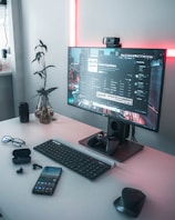 A modern workspace featuring a desktop computer with a large monitor displaying a playlist and music application. Surrounding the monitor are various electronic devices including a smartphone, wireless earphones, a keyboard, a mouse, and a gaming controller. A potted plant adds a natural element, and soft pink lighting provides a warm ambiance.