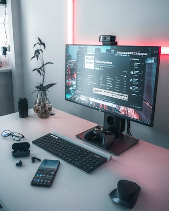 A modern workspace featuring a desktop computer with a large monitor displaying a playlist and music application. Surrounding the monitor are various electronic devices including a smartphone, wireless earphones, a keyboard, a mouse, and a gaming controller. A potted plant adds a natural element, and soft pink lighting provides a warm ambiance.