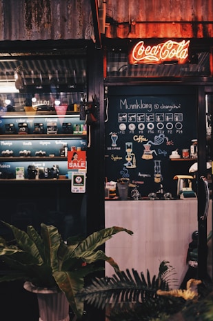 A vintage-style coffee shop with a neon Coca-Cola sign above the entrance. The interior features a chalkboard menu with various coffee options, and there is an assortment of cameras displayed on shelves. A sale sign is placed near the door. Large potted plants add greenery to the scene.