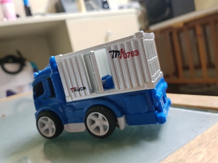 A toy truck with a blue cab and a white cargo container featuring the numbers 0783 and the word TRUCK. It rests on a glass surface with a blurred indoor background, including shelves and a wooden object.
