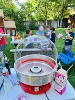 A backyard setting with a group of children gathered around a table, enjoying cotton candy. A red cotton candy machine is in the foreground, and various items like flossugar, bottles, and paper towels are on the table. Lush greenery and a wooden playhouse are visible in the background, creating a playful and lively atmosphere.