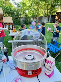 A backyard setting with a group of children gathered around a table, enjoying cotton candy. A red cotton candy machine is in the foreground, and various items like flossugar, bottles, and paper towels are on the table. Lush greenery and a wooden playhouse are visible in the background, creating a playful and lively atmosphere.