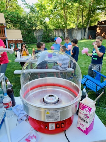 A backyard setting with a group of children gathered around a table, enjoying cotton candy. A red cotton candy machine is in the foreground, and various items like flossugar, bottles, and paper towels are on the table. Lush greenery and a wooden playhouse are visible in the background, creating a playful and lively atmosphere.