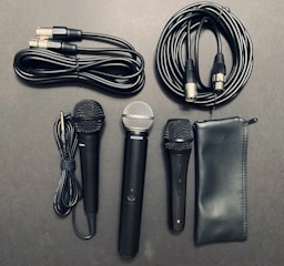 A collection of audio equipment including three microphones of different designs, several audio cables, and a black zippered pouch. The microphones feature both silver and black grills, and the cables have various connectors like XLR and quarter-inch jacks.
