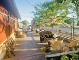 A sunlit outdoor patio with multiple wicker chairs and couches arranged around tables. String lights hang above, creating a cozy atmosphere. The area is bordered by a metal fence and surrounded by lush green trees. Wooden barrels and potted plants add decorative elements to the setting.