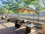 An outdoor seating area is set in a shaded patio with large umbrellas, surrounded by lush green trees and enclosed by a metal fence. The space includes wicker chairs, black cushioned sofas, and a coffee table made from a vintage trunk, creating an inviting and comfortable atmosphere.