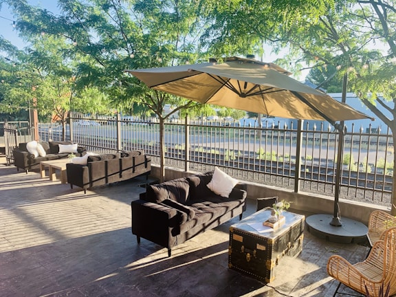 An outdoor seating area is set in a shaded patio with large umbrellas, surrounded by lush green trees and enclosed by a metal fence. The space includes wicker chairs, black cushioned sofas, and a coffee table made from a vintage trunk, creating an inviting and comfortable atmosphere.