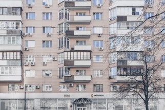 An apartment building with a beige facade features multiple windows, some with balconies enclosed by glass. Air conditioning units are mounted beneath several windows. The lower level has commercial establishments, including a visible post office sign. Leafless trees partially obscure parts of the building.