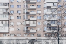 An apartment building with a beige facade features multiple windows, some with balconies enclosed by glass. Air conditioning units are mounted beneath several windows. The lower level has commercial establishments, including a visible post office sign. Leafless trees partially obscure parts of the building.