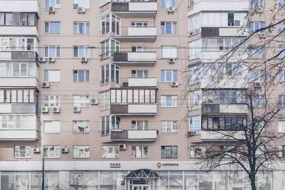 An apartment building with a beige facade features multiple windows, some with balconies enclosed by glass. Air conditioning units are mounted beneath several windows. The lower level has commercial establishments, including a visible post office sign. Leafless trees partially obscure parts of the building.