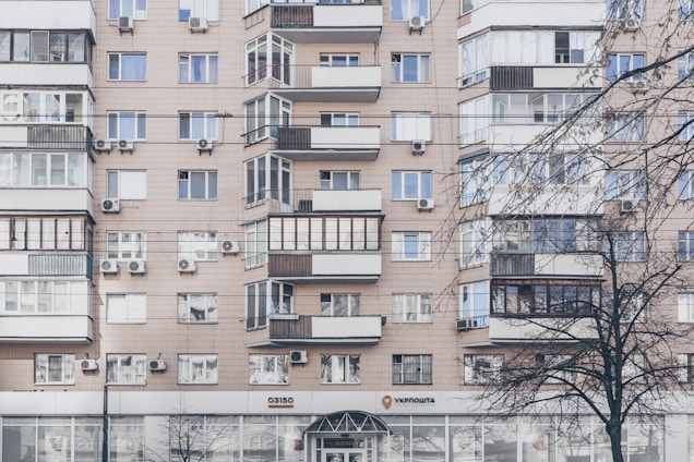 An apartment building with a beige facade features multiple windows, some with balconies enclosed by glass. Air conditioning units are mounted beneath several windows. The lower level has commercial establishments, including a visible post office sign. Leafless trees partially obscure parts of the building.