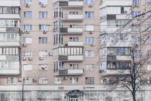 An apartment building with a beige facade features multiple windows, some with balconies enclosed by glass. Air conditioning units are mounted beneath several windows. The lower level has commercial establishments, including a visible post office sign. Leafless trees partially obscure parts of the building.